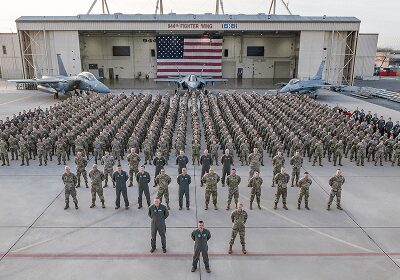 Large group of military personnel standing in formation with large American flag.